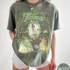 May include: Moss green t-shirt with a framed illustration of Princess Tiana. The design includes the name "Tiana" in green script, a portrait of Tiana, and characters from The Princess and the Frog. The shirt has a relaxed fit and is a moss green colour.