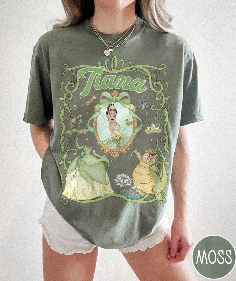May include: Moss green t-shirt with a framed illustration of Princess Tiana. The design includes the name "Tiana" in green script, a portrait of Tiana, and characters from The Princess and the Frog. The shirt has a relaxed fit and is a moss green colour.