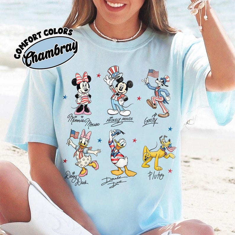 May include: Pale blue t-shirt with a graphic of Mickey Mouse, Minnie Mouse, Goofy, Daisy Duck, Donald Duck, and Pluto in patriotic outfits. The shirt has the words "COMFORT COLORS" and "Chambray" printed on it.