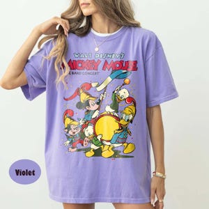 May include: Violet-colored t-shirt featuring a graphic of Mickey Mouse and friends in a band concert. The text "WALT DISNEY'S MICKEY MOUSE BAND CONCERT" is printed across the top. The shirt has short sleeves and a crew neck.