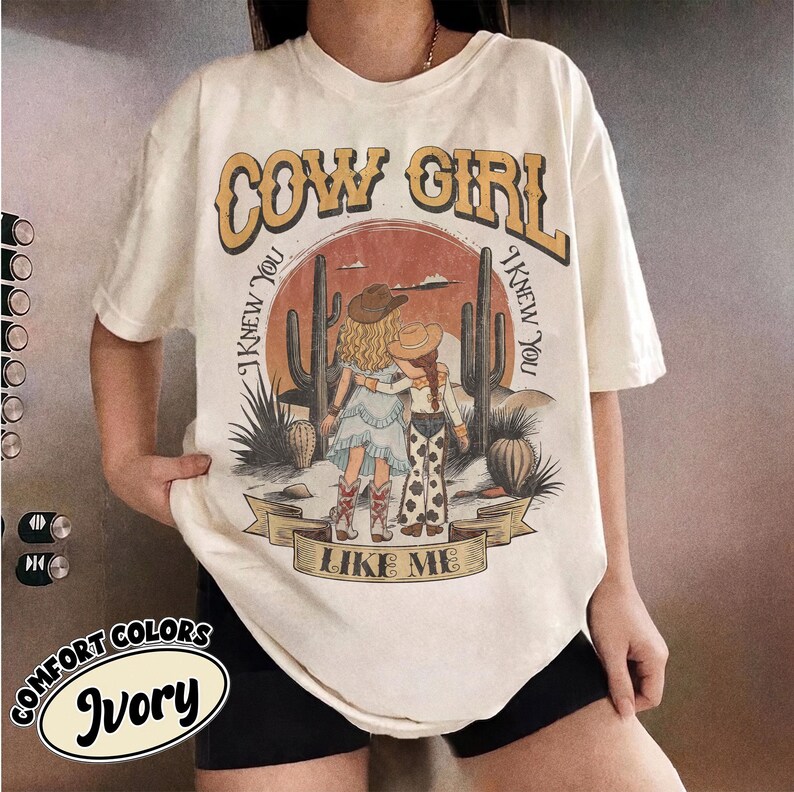 May include: An ivory short-sleeve t-shirt featuring a graphic print of two figures in cowboy hats standing in a desert landscape with cacti. The text reads COW GIRL at the top and I KNEW YOU I KNEW YOU LIKE ME around the central illustration.