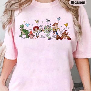 May include: A pale pink t-shirt with a cartoon design of Toy Story characters. The graphic includes Rex, Hamm, Jessie, Buzz Lightyear, Woody, and Bullseye, each with their names underneath.