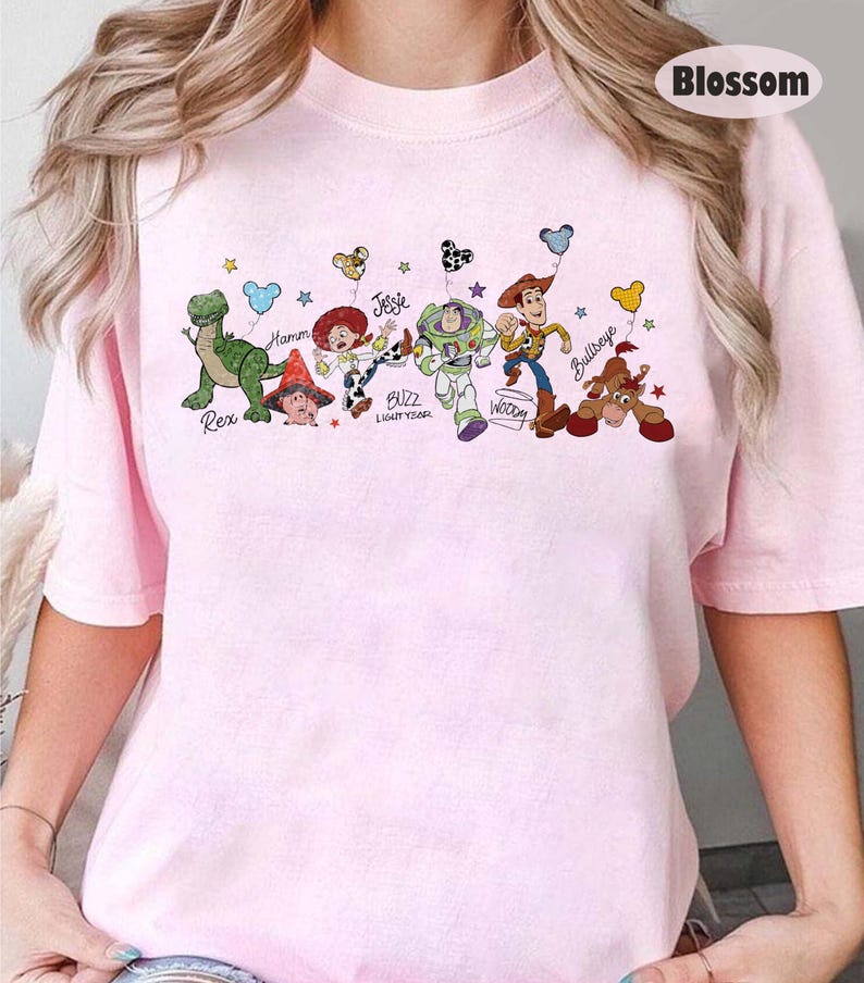 May include: A pale pink t-shirt with a cartoon design of Toy Story characters. The graphic includes Rex, Hamm, Jessie, Buzz Lightyear, Woody, and Bullseye, each with their names underneath.