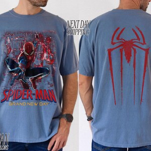 以下が含まれることがあります： スパイダーマンが描かれたブルージーンズのTシャツ。前面には、スパイダーマンがアクションを起こしているグラフィックと「Spider-Man Brand New Day」の文字が描かれています。Tシャツの背面には赤いクモのシンボルが表示されています。