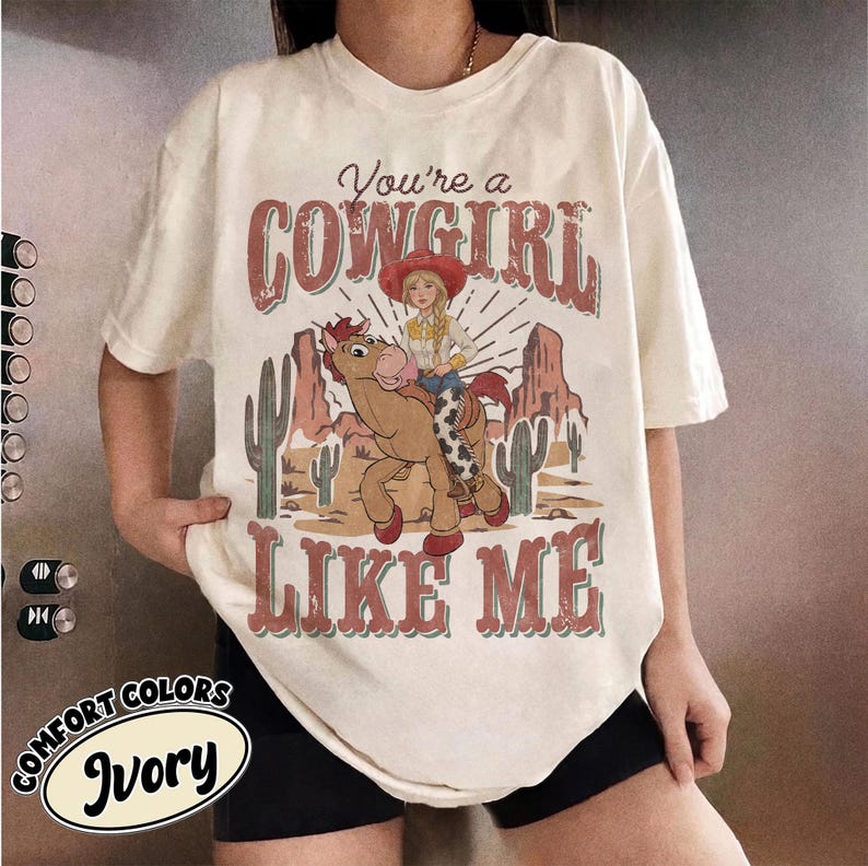 May include: An ivory t-shirt featuring a graphic print of a cartoon cowgirl riding a horse in a desert landscape with cacti and rock formations. The text reads "You're a COWGIRL LIKE ME" in a distressed red font with a thin teal outline.