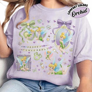 May include: A light purple t-shirt featuring multiple illustrations of Tinker Bell in various poses, surrounded by small hearts, butterflies, and gems. The text "DISNEY TINKERBELL" is printed in the center, with a "COMFORT COLORS Orchid" label.