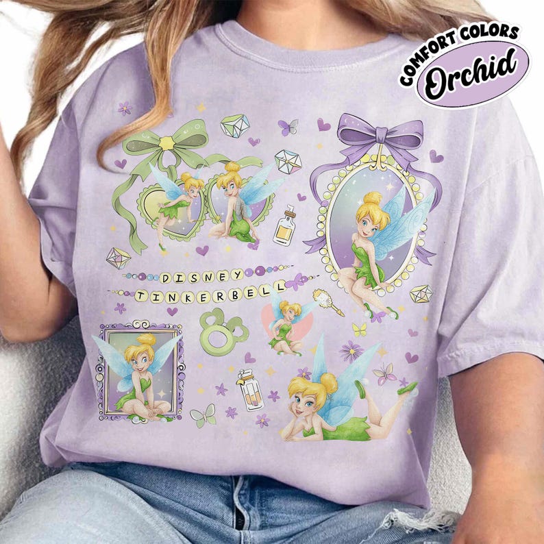 May include: A light purple t-shirt featuring multiple illustrations of Tinker Bell in various poses, surrounded by small hearts, butterflies, and gems. The text "DISNEY TINKERBELL" is printed in the center, with a "COMFORT COLORS Orchid" label.