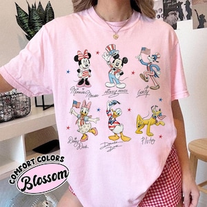 May include: A light pink Comfort Colors t-shirt with cartoon characters Minnie Mouse, Mickey Mouse, Goofy, Daisy Duck, Donald Duck, and Pluto in patriotic outfits. The shirt includes the text "Comfort Colors Blossom".