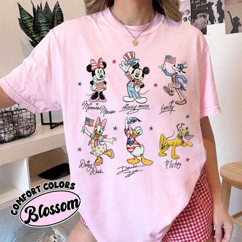 May include: A light pink Comfort Colors t-shirt with cartoon characters Minnie Mouse, Mickey Mouse, Goofy, Daisy Duck, Donald Duck, and Pluto in patriotic outfits. The shirt includes the text "Comfort Colors Blossom".