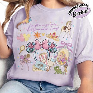 May include: Orchid-colored t-shirt with a graphic featuring Disney-themed illustrations. The design includes a princess, a horse, a crown, and Mickey Mouse ears. Text on the shirt reads "I've got a magic hair that glows when I sing!" and "Let the Magic Begin!"