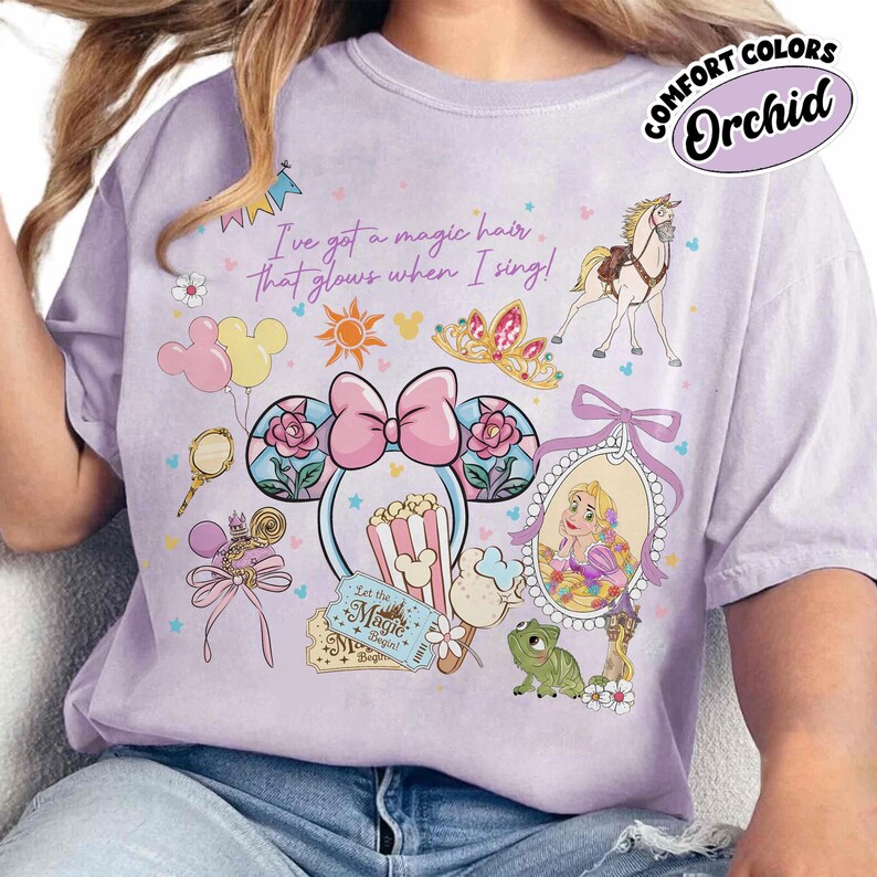 May include: Orchid-colored t-shirt with a graphic featuring Disney-themed illustrations. The design includes a princess, a horse, a crown, and Mickey Mouse ears. Text on the shirt reads "I've got a magic hair that glows when I sing!" and "Let the Magic Begin!"