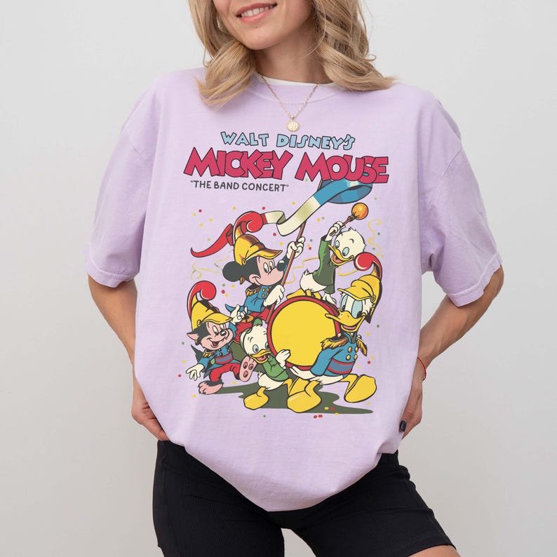 May include: A light purple t-shirt featuring a graphic of Mickey Mouse and friends in a band concert. The text "Walt Disney's Mickey Mouse" is above the image. The band includes Mickey, Donald, Goofy, and other characters.