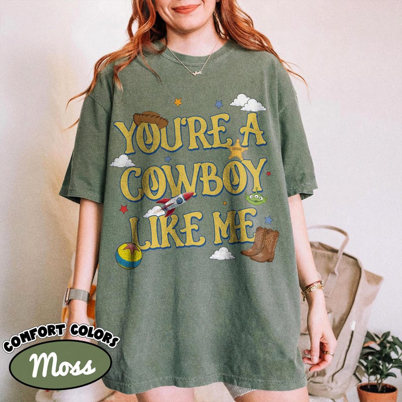 Puede incluir: Una camiseta de manga corta color verde musgo con el texto amarillo "YOU'RE A COWBOY LIKE ME" e ilustraciones gr&aacute;ficas de un sombrero de vaquero marr&oacute;n, un cohete, una pelota colorida, un alien&iacute;gena verde y botas vaqueras marrones.