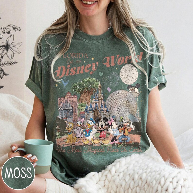 K&ouml;nnte beinhalten: Moosgr&uuml;nes T-Shirt mit einem Retro-Grafikdesign von Disney World in Florida. Das Design zeigt ber&uuml;hmte Wahrzeichen, Feuerwerk und Charaktere wie Mickey Mouse und Donald Duck. Der Text lautet "Florida Disney World" und "The Most Magical Place on Earth."