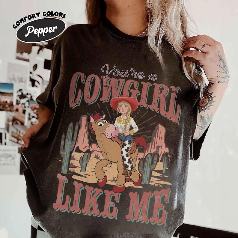 May include: A pepper-coloured t-shirt featuring a graphic of a blonde cowgirl riding a brown horse in a desert landscape with cacti. The text reads You're a COWGIRL LIKE ME in red and teal lettering. The shirt has a crew neck and short sleeves.