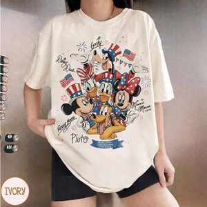 May include: Ivory t-shirt with a graphic of Mickey Mouse, Minnie Mouse, Donald Duck, Daisy Duck, Goofy, and Pluto in patriotic outfits. The design includes American flags and fireworks, with text that reads "Mickey Mouse", "Pluto", and "USA Anniversary 1776-2023".