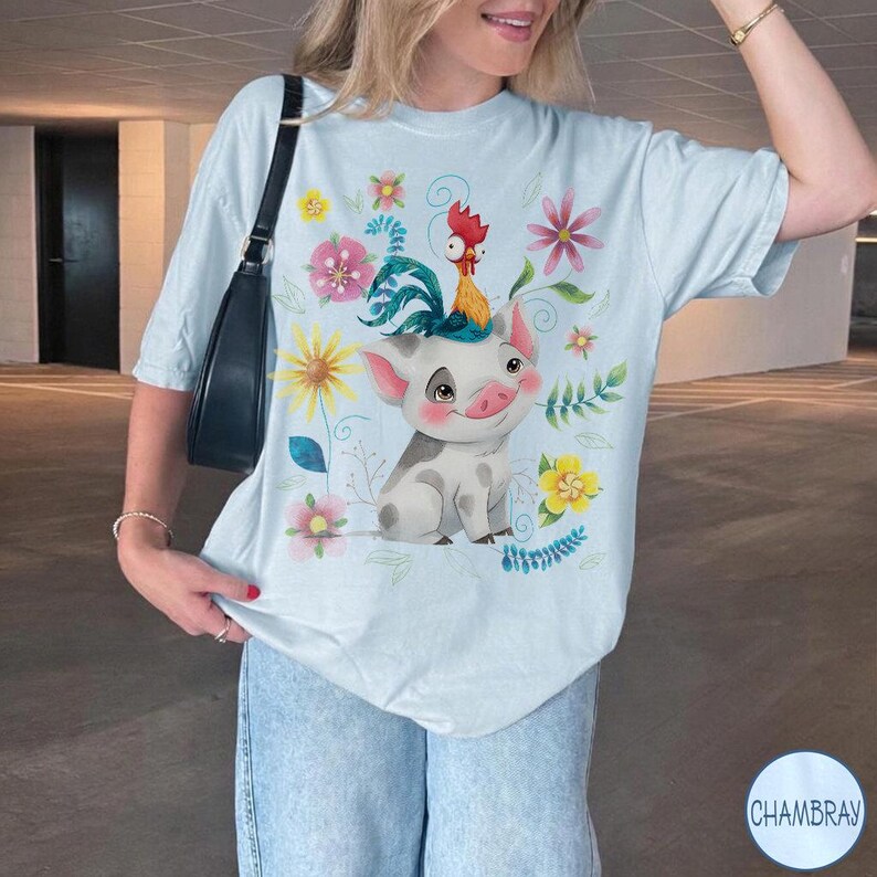 Pu&ograve; includere: T-shirt azzurra con un disegno di maiale e gallo dei cartoni animati, circondato da fiori colorati. Il design include elementi floreali gialli, rosa e blu. La maglietta ha una vestibilit&agrave; rilassata.