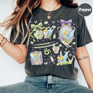 May include: A charcoal gray t-shirt featuring multiple illustrations of Tinkerbell in various poses, surrounded by decorative frames, bows, butterflies, and small gems. The text Disney Tinkerbell is printed in the center with a small bottle icon.