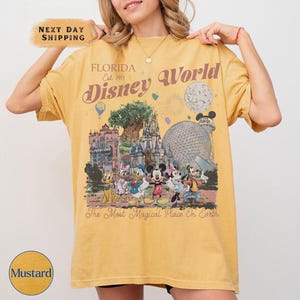 K&ouml;nnte beinhalten: Senffarbenes T-Shirt mit einem Disney World-Motiv im Vintage-Stil. Das Design enth&auml;lt den Text "FLORIDA Est. 1971 Disney World" und Disney-Figuren. Der Satz "The Most Magical Place On Earth" ist ebenfalls vorhanden.