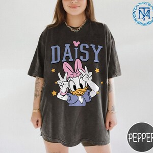 Peut inclure: T-shirt gris fonc&eacute; avec un motif de Daisy Duck avec un n&oelig;ud rose et le mot "DAISY" en bleu. Des &eacute;toiles jaunes entourent le personnage. Le t-shirt a des manches courtes et une coupe ample.