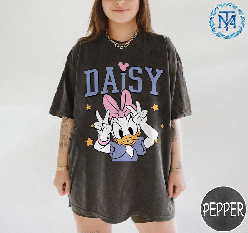 Peut inclure: T-shirt gris fonc&eacute; avec un motif de Daisy Duck avec un n&oelig;ud rose et le mot "DAISY" en bleu. Des &eacute;toiles jaunes entourent le personnage. Le t-shirt a des manches courtes et une coupe ample.