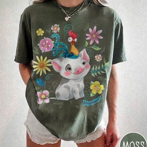 Pu&ograve; includere: T-shirt verde muschio con un design a fumetto di maiale e gallo, circondato da fiori e foglie colorate. L'illustrazione ha uno stile allegro e stravagante. La maglietta ha una vestibilit&agrave; ampia e maniche corte.