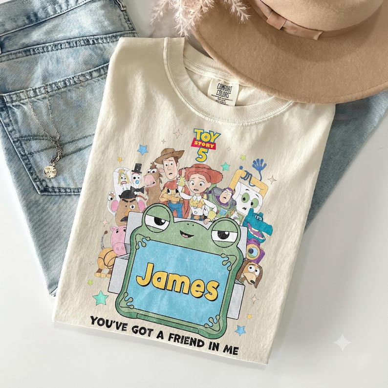 May include: A cream-colored t-shirt featuring a graphic print of Toy Story 5 characters surrounding a green frog-shaped frame with the name James in yellow text. The shirt includes the text Toy Story 5 and You've Got A Friend In Me.