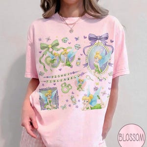 May include: A light pink short-sleeve t-shirt featuring multiple illustrations of Tinker Bell, green bows, purple frames, and small gems. The text "DISNEY TINKERBELL" is printed in the center. The design includes various butterflies and floral accents.