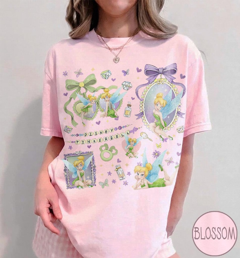 May include: A light pink short-sleeve t-shirt featuring multiple illustrations of Tinker Bell, green bows, purple frames, and small gems. The text "DISNEY TINKERBELL" is printed in the center. The design includes various butterflies and floral accents.