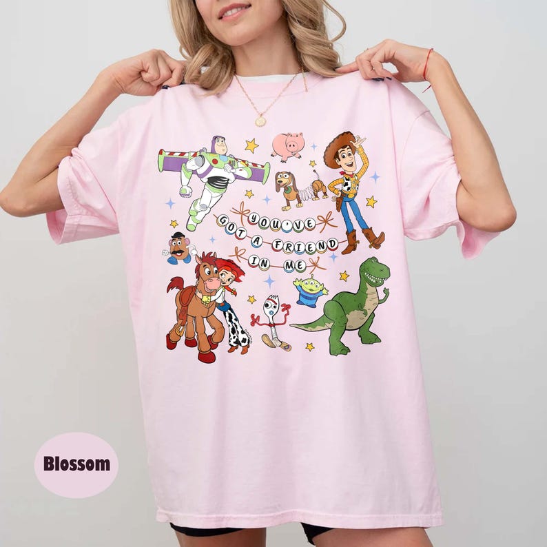 May include: A pale pink t-shirt with a vibrant graphic of animated characters and the text "YOU'VE GOT A FRIEND IN ME". The design includes a space ranger, a cowboy, a dinosaur, and other characters.