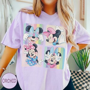 May include: A light purple t-shirt featuring four squares with Minnie Mouse illustrations. Each Minnie Mouse has a different pose and bow color, set against a pastel rainbow background. The word "ORCHID" is printed on the bottom left.