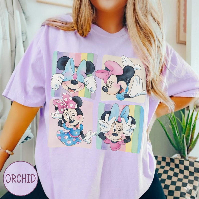 May include: A light purple t-shirt featuring four squares with Minnie Mouse illustrations. Each Minnie Mouse has a different pose and bow color, set against a pastel rainbow background. The word "ORCHID" is printed on the bottom left.