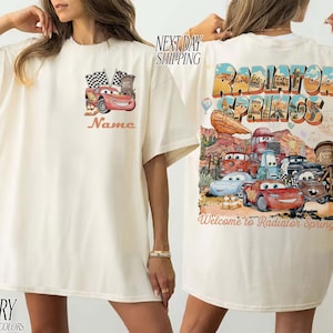 Puede incluir: Camiseta de color marfil con un gr&aacute;fico de los personajes de la pel&iacute;cula Cars. La parte delantera presenta a Rayo McQueen con banderas a cuadros y la palabra "Name". La parte trasera muestra "Radiator Springs" y "Welcome to Radiator Springs".