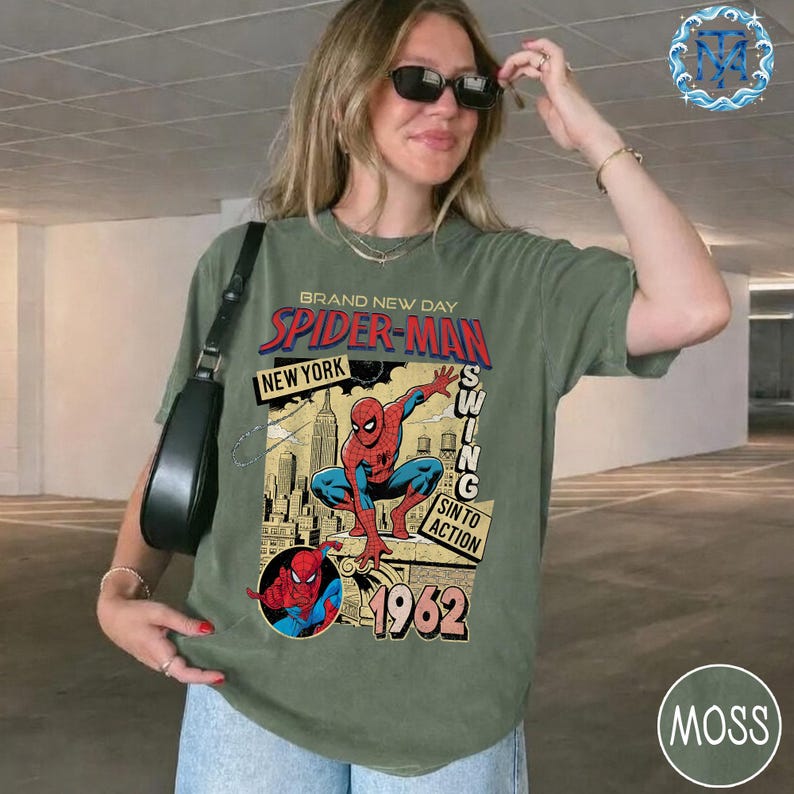 May include: Olive green t-shirt featuring a Spider-Man graphic. The design includes the text "BRAND NEW DAY SPIDER-MAN NEW YORK" with an illustration of Spider-Man and the year "1962". The shirt has a vintage, distressed look.