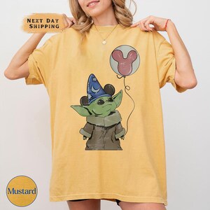 Puede incluir: Camiseta de color mostaza con un gr&aacute;fico de un personaje alien&iacute;gena verde que lleva un sombrero de mago azul y sostiene un globo de Mickey Mouse. La camiseta tiene el texto "Next Day Shipping" y "Mustard".
