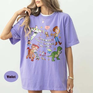 May include: A violet t-shirt with a graphic print of characters from the film Toy Story. The design includes Buzz Lightyear, Woody, and other characters, with the phrase "You've Got a Friend in Me."