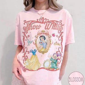 May include: Pale pink t-shirt with a Snow White design. The graphic includes an oval portrait of Snow White, her dress, a dwarf, and the words "Snow White" in a decorative frame. A crown, apple, and bird are also included.