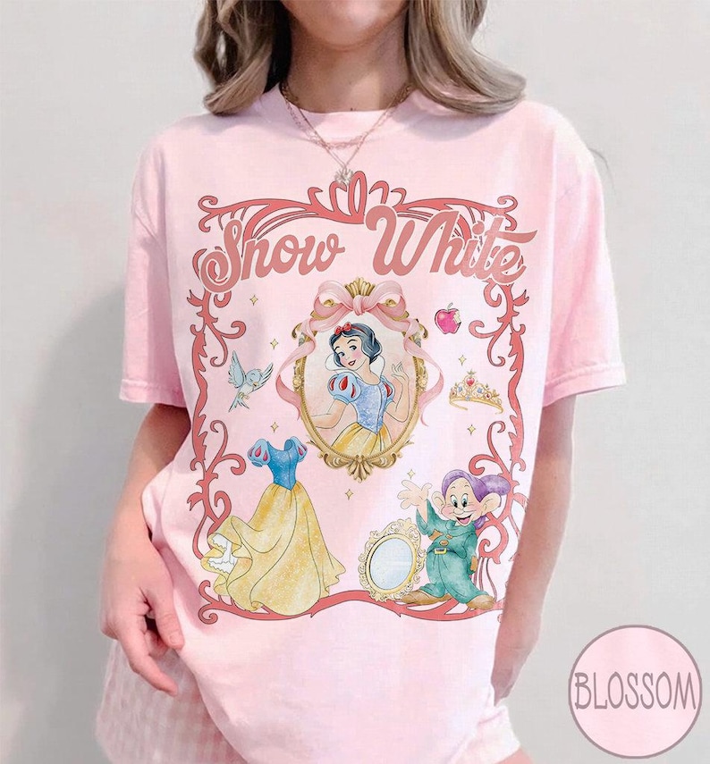 May include: Pale pink t-shirt with a Snow White design. The graphic includes an oval portrait of Snow White, her dress, a dwarf, and the words "Snow White" in a decorative frame. A crown, apple, and bird are also included.
