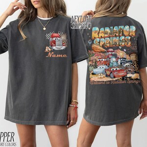 Puede incluir: Camiseta gris oscuro con un cami&oacute;n de dibujos animados, una bandera a cuadros y la palabra "Name" en naranja. La parte trasera muestra un gr&aacute;fico colorido de "Radiator Springs" con personajes de coches y el texto "Welcome to Radiator Springs."