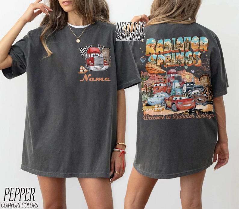 Puede incluir: Camiseta gris oscuro con un cami&oacute;n de dibujos animados, una bandera a cuadros y la palabra "Name" en naranja. La parte trasera muestra un gr&aacute;fico colorido de "Radiator Springs" con personajes de coches y el texto "Welcome to Radiator Springs."