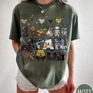 May include: An olive green Star Wars t-shirt with cartoon character illustrations and the Star Wars logo. The design includes Darth Vader, Chewbacca, Grogu, BB-8, a Stormtrooper, and R2-D2. The shirt has a crew neck and short sleeves.