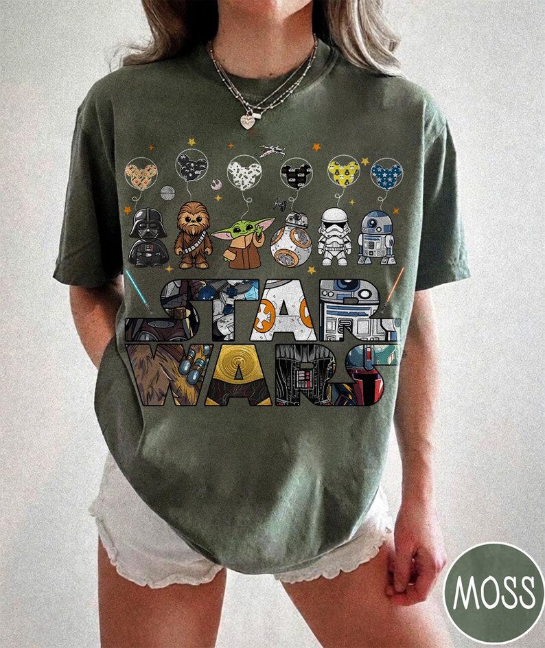 May include: An olive green Star Wars t-shirt with cartoon character illustrations and the Star Wars logo. The design includes Darth Vader, Chewbacca, Grogu, BB-8, a Stormtrooper, and R2-D2. The shirt has a crew neck and short sleeves.