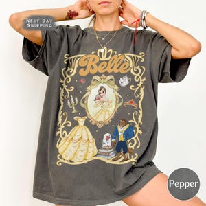 May include: Dark grey t-shirt with a Belle graphic. The design includes an illustration of Belle in a yellow dress, the Beast, and other elements from the movie. The text "Belle" is displayed in a retro font. The shirt also has the text "Next Day Shipping".