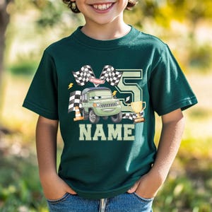 Pode incluir: T-shirt verde escuro com um carro de desenho animado, bandeiras quadriculadas, um trof&eacute;u e o n&uacute;mero 5. A palavra "NAME" &eacute; impressa em letras brancas grandes abaixo do gr&aacute;fico do carro. Um design divertido para o anivers&aacute;rio de uma crian&ccedil;a.