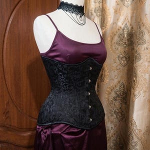May include: A dark purple satin camisole top paired with a black brocade corset featuring a front busk closure with silver-toned metal clasps. A black lace choker with delicate hanging chains is worn around the neck of the mannequin display.