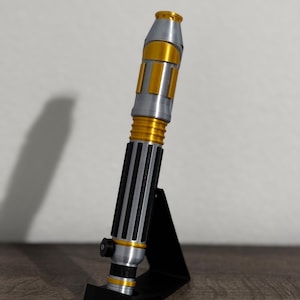 Peut inclure: Un accessoire de sabre laser argenté et doré, avec une poignée noire à rainures verticales. L'accessoire est présenté sur un support noir. Le sabre laser a des accents dorés sur le dessus, le milieu et le bas.