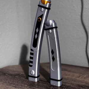 Pu&ograve; includere: Due spade laser argentate con dettagli neri e dorati. La spada laser pi&ugrave; grande presenta un pannello nero con diamanti e scanalature. La spada laser pi&ugrave; piccola ha un design simile. Entrambe hanno fasce nere e sono esposte su una superficie di legno.
