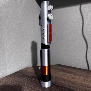 May include: A silver and black lightsaber prop with red and orange accents. The prop has a detailed design with buttons and a rectangular red panel. The lightsaber is displayed on a wooden surface.