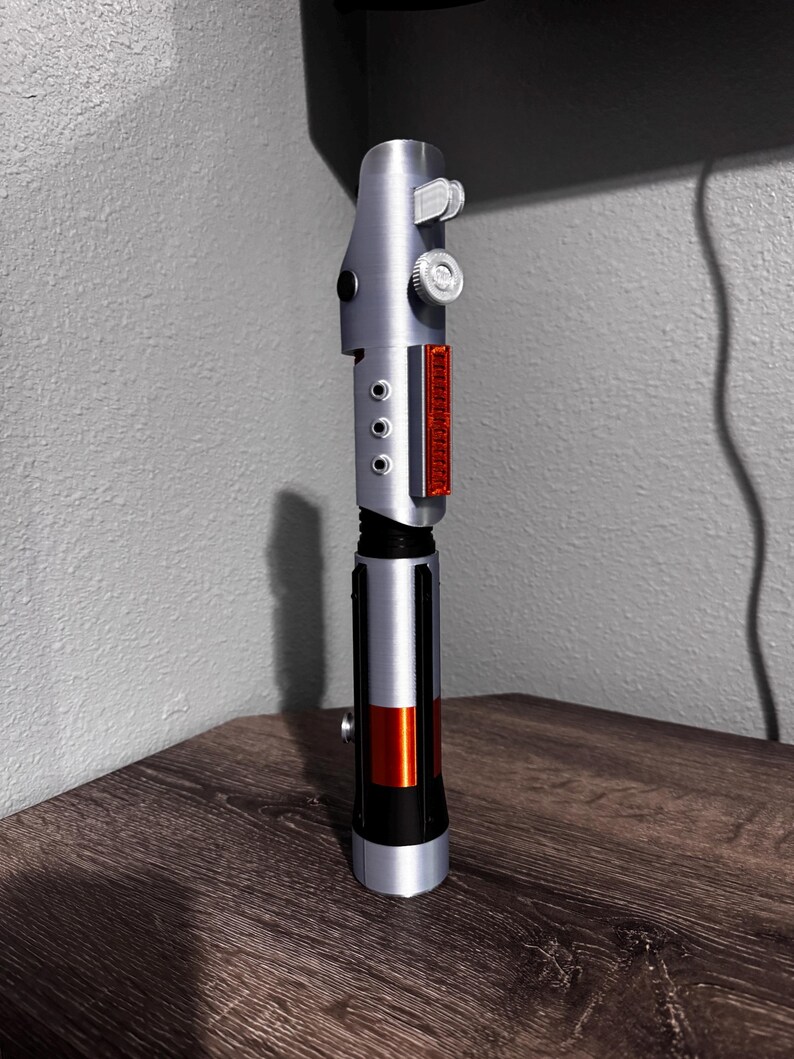 May include: A silver and black lightsaber prop with red and orange accents. The prop has a detailed design with buttons and a rectangular red panel. The lightsaber is displayed on a wooden surface.