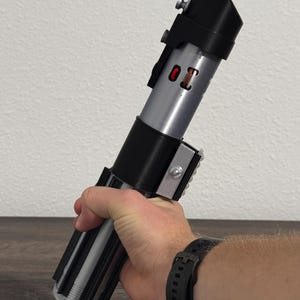 Pu&ograve; includere: Un'elica di spada laser argento e nera con un pulsante rosso e la scritta "01". L'elica ha un'impugnatura nera e un corpo argentato. La spada laser &egrave; tenuta in mano.
